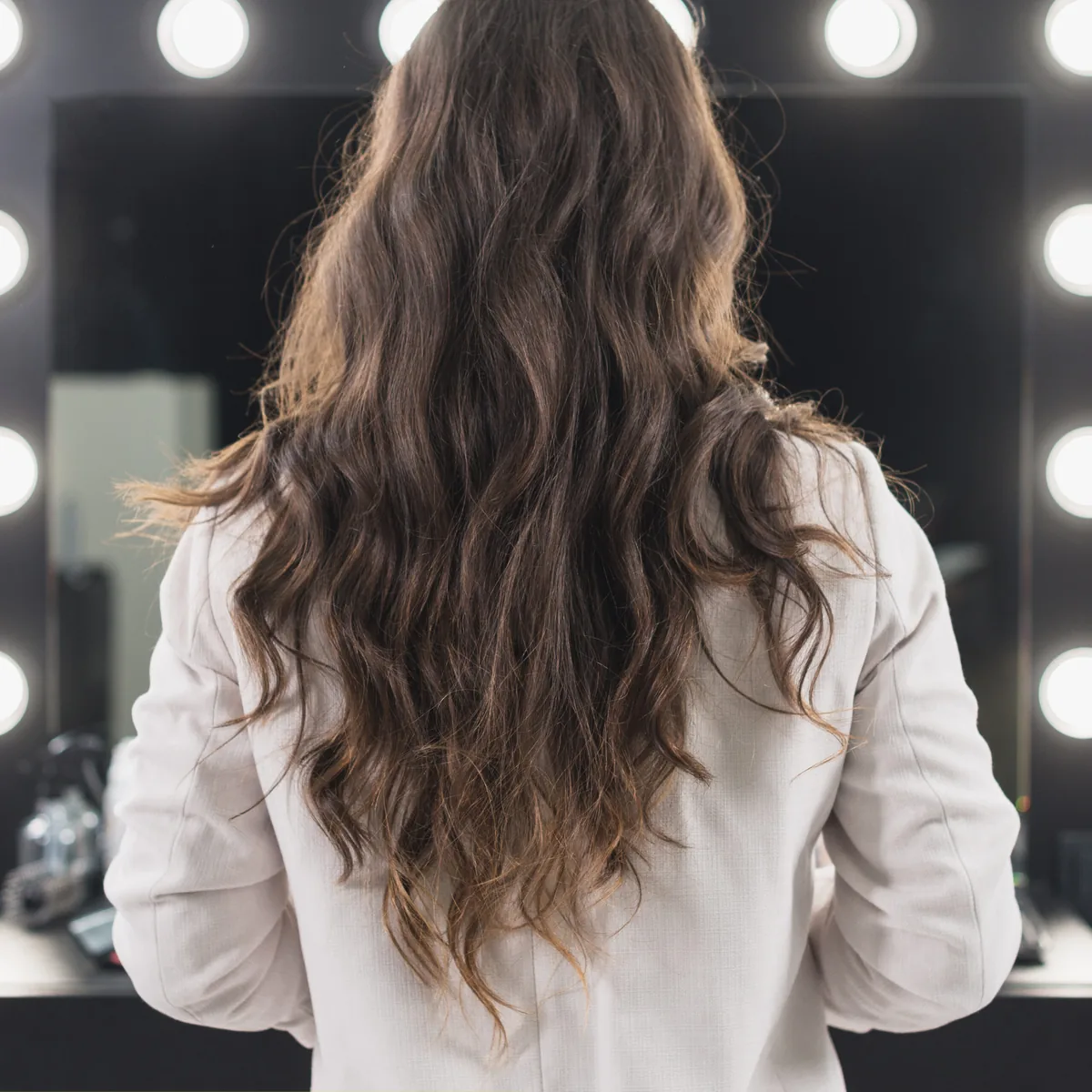 Brunette hand tied extensions seen from behind in front of a bulb lit vanity mirror.